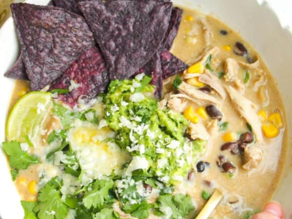 White chicken chili in a white bowl, topped with avocado, cheese and tortilla chips.