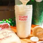 Kewpie mayo in a tall glass jar on a wooden board with a spoon on top.