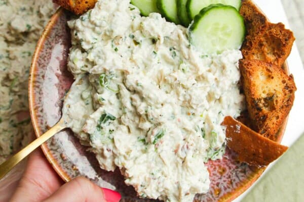 Hand holding plate with Caesar inspired chicken salad, with pita chips and cucumber on the side.